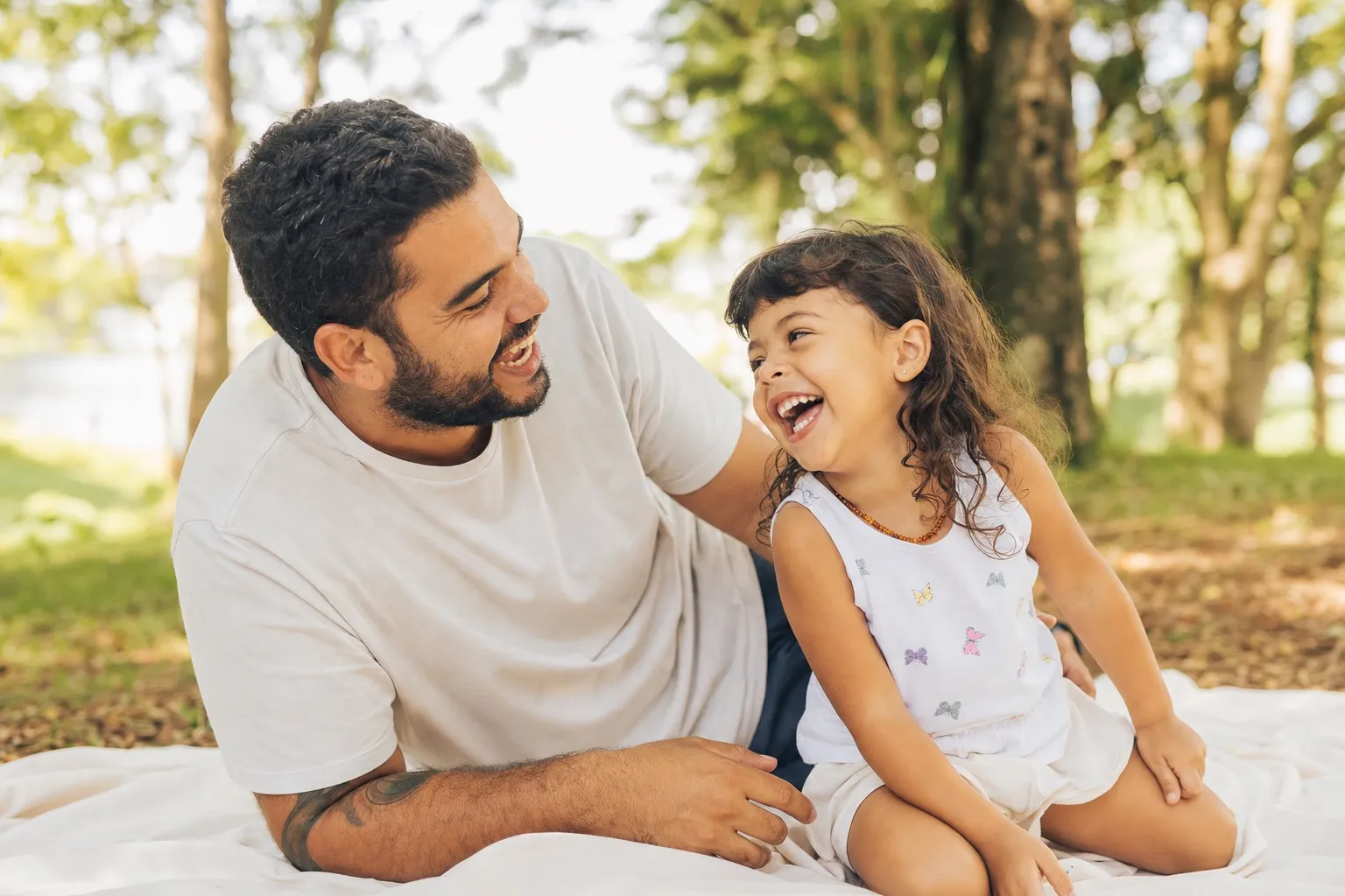 Padre e hija pequeña riendo juntos sobre una manta en un picnic al aire libre, con luz natural de tarde.