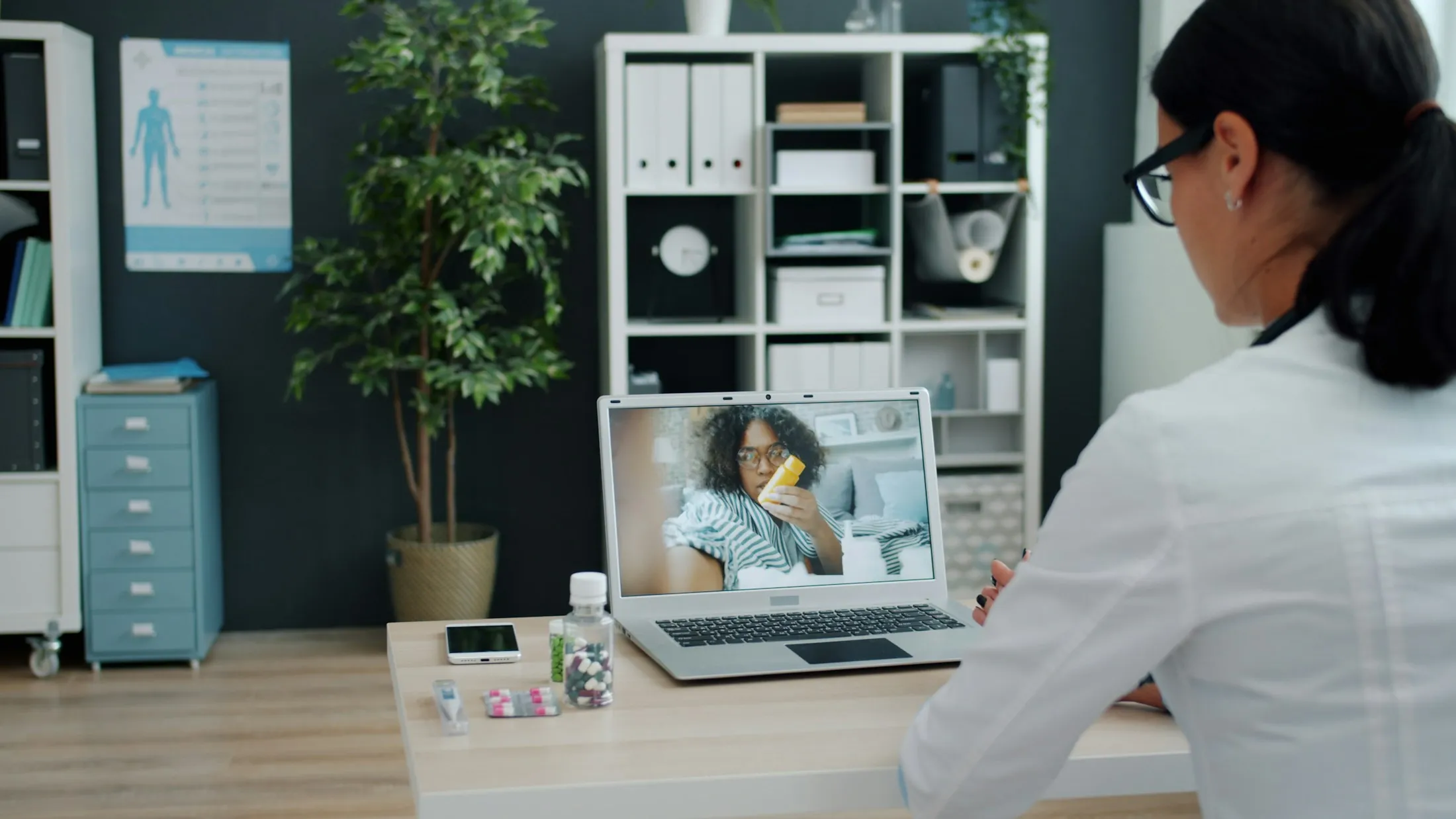 Profesional de salud con bata blanca en una videollamada desde una laptop, escritorio de oficina con luz natural, estantería y planta al fondo.